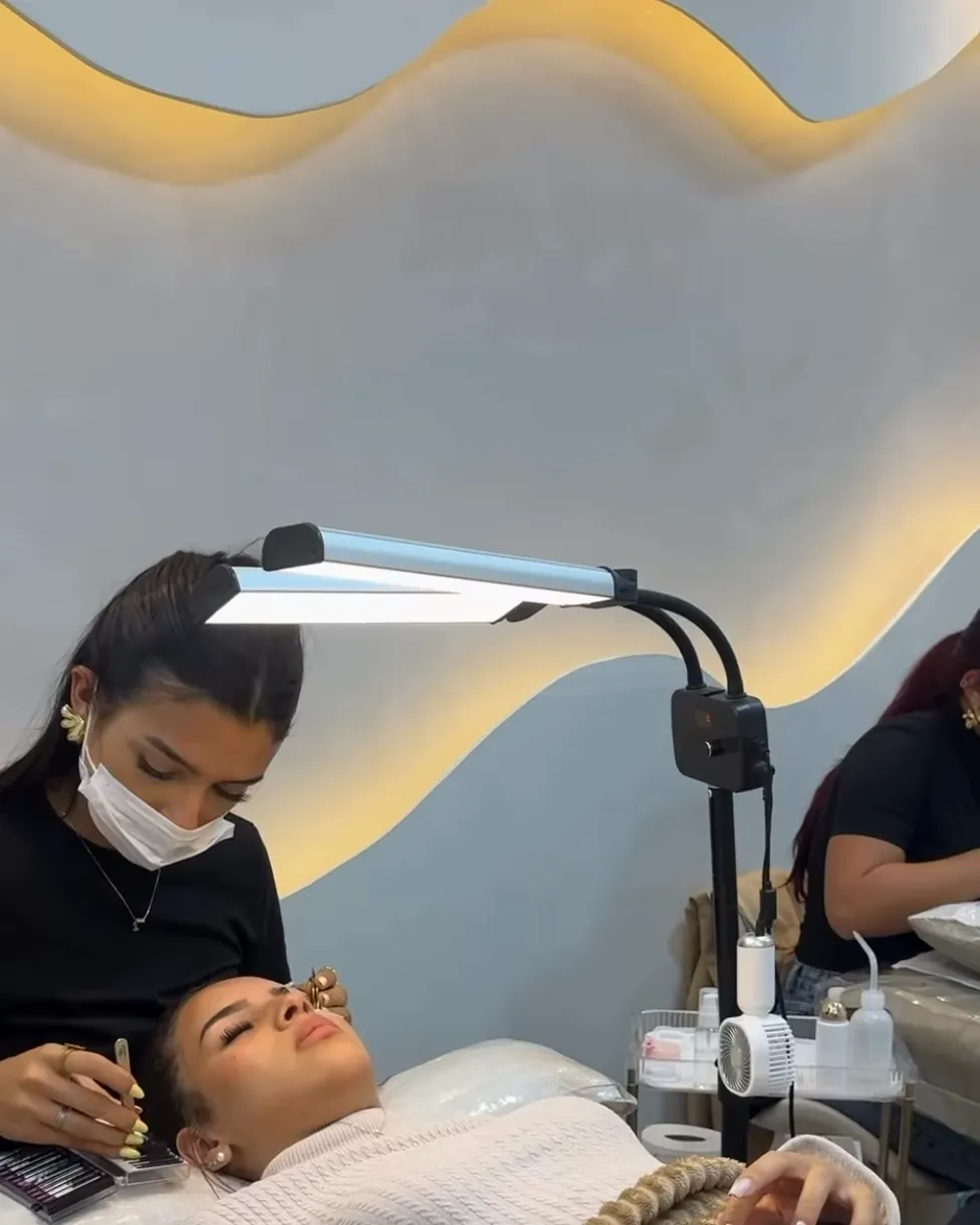Profissional do studio aplicando cílios numa cliente deitada na maca, sob a parede iluminada em ondas